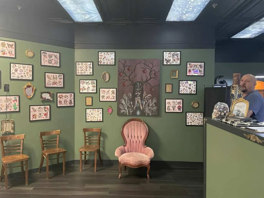 The waiting room at Legacy Tattoo Company, with sage green walls covered in framed flash sheets, wooden chairs, and a blush velvet armchair